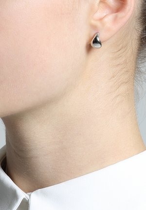 Clou d'oreille en forme de goutte d'argent porté en gros plan sur l'oreille gauche et le cou avec une chemise blanche à col.