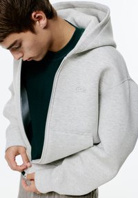 Svetlo šedá mikina na zips s kapucňou na šnúrku a prednými vreckami, vrstvená nad tmavozeleným tričkom. Má priestraný, uvoľnený strih.