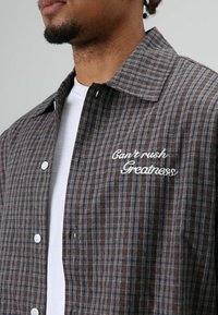 Bruin geruit overhemd met een standaardkraag, knoopsluiting en witte geborduurde tekst "Can't rush Greatness" op de linkerborst.