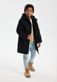 Veste doudoune noire avec capuche, tissu texturé et poches avant. Portée sur un pull beige et associée à un jean bleu clair et des bottes beige.