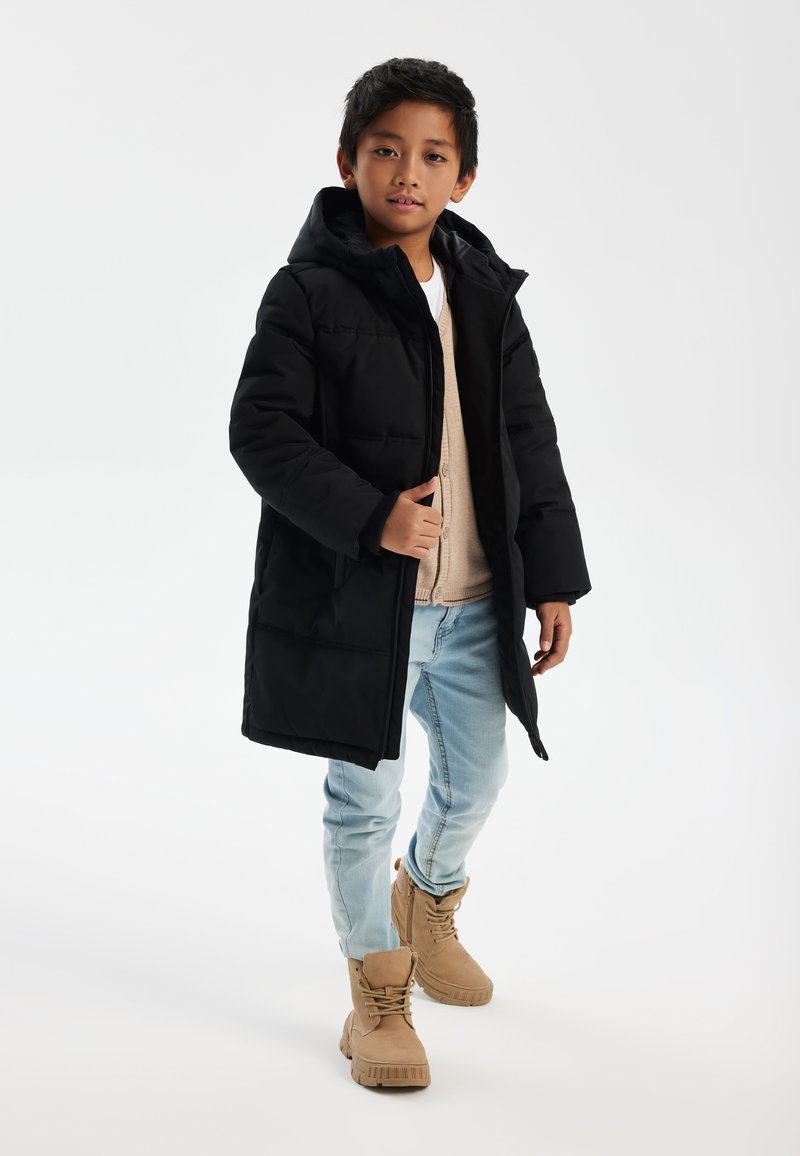 Veste doudoune noire avec capuche, tissu texturé et poches avant. Portée sur un pull beige et associée à un jean bleu clair et des bottes beige.