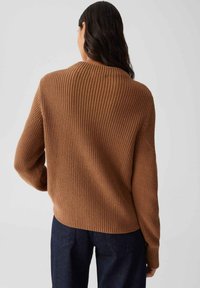 Frau mit langen, dunklen Haaren, von hinten gezeigt, trägt einen braunen, gerippten Pullover und dunkelblaue Jeans vor einem schlichten Hintergrund.