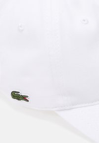 Hvit bomullslue med teksturert stoff. Har en grønn brodert krokodillelogo på siden. Buet brem og sydd detaljer.