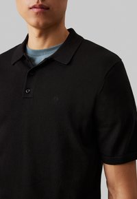 Polo nero con tessuto strutturato, maniche corte e colletto. Presenta tre bottoni e un piccolo logo in rilievo sul lato sinistro del petto.