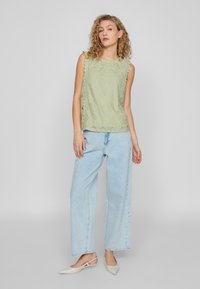Top in pizzo verde chiaro con bordi scallop, abbinato a pantaloni in denim chiaro e larghi. Le scarpe presentano una punta appuntita e una cinghia alla caviglia.