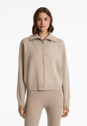 Beige Reißverschluss-Pullover mit Kragen, gerippter Textur und überschnittenen Schultern. Kombiniert mit passenden beigen, figurbetonten Hosen für einen stimmigen Look.