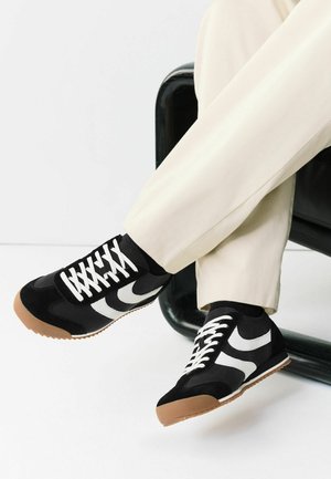 Person wearing black and white lace-up sneakers with gum soles and cream-colored pants, sitting with legs crossed on a black chair.