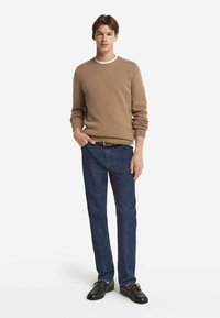 Jasnobrązowy sweter dzianinowy z ściągaczami na rękawach, zestawiony z ciemnoniebieskimi spodniami typu straight-leg. Model nosi ciemne buty z brązowymi sznurowadłami dla kontrastu.