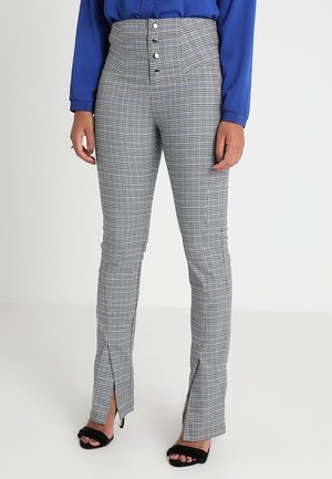 Femme portant un pantalon taille haute à carreaux noirs et blancs avec des boutons à l'avant et des fentes à l'ourlet avant, associé à des talons ouverts noirs et un chemisier bleu.