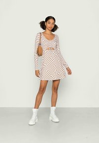 Vestido de manga longa em xadrez castanho e branco com detalhes em recorte, saia evasê e botas brancas combinando. Acessório com uma bolsa castanha.