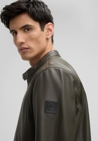 Olivgrüne Lederjacke mit glatter Textur, ausgestattet mit einem Stehkragen und einem schwarzen Logo-Patch auf dem linken Ärmel.