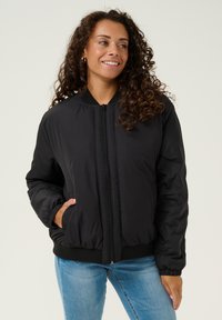 Giubbotto bomber nero realizzato in nylon liscio, con zip frontale, polsini e orlo a coste, e tasche laterali. Abbinato a jeans azzurri chiari.