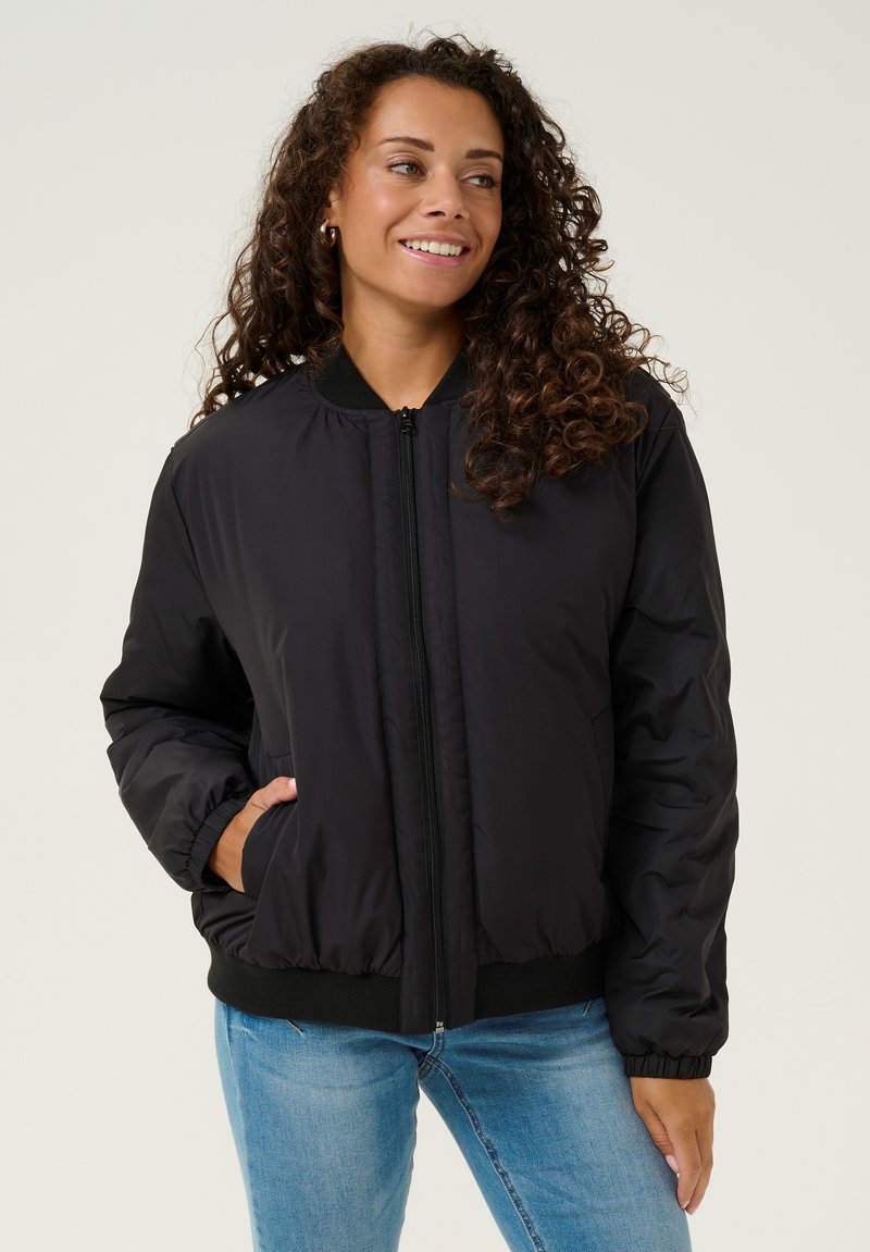 Giubbotto bomber nero realizzato in nylon liscio, con zip frontale, polsini e orlo a coste, e tasche laterali. Abbinato a jeans azzurri chiari.