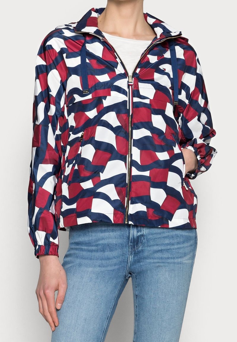 Bunda větrovka s odvážným vzorem v červené, bílé a námořnické modři, se zapínáním na zip, nastavovacími šňůrkami a elastickými manžetami. Vyrobena z lehkého materiálu.