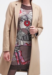 Abrigo beige ajustado sobre un vestido estampado que presenta un diseño gráfico con un motivo floral y de personajes en colores marrón, rojo y negro.