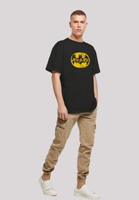 Black t-shirt with a yellow Batman logo and Japanese text. Model wears beige cargo pants and white shoes. Short sleeves, relaxed fit.