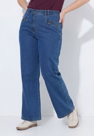 Person trägt lockere blaue Denim-Jeans mit vorderen Reißverschlusstaschen und weißen Sneakers, steht vor einem schlichten hellen Hintergrund.