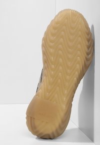 Sula till sko med ett vågigt mönster, tillverkad av lätt gummi. Färgen är beige med en texturerad yta. Har varumärkeslogotyp nära hälen.