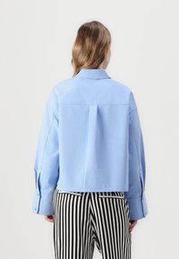 Camicia corta azzurro chiaro realizzata in tessuto dalla texture liscia. Presenta un colletto e polsini sbottonati, indossata con pantaloni a righe bianche e nere.