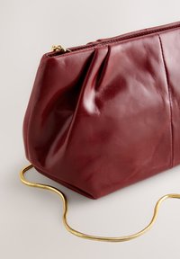 Bordeauxrode leren clutch met een geplooid ontwerp, voorzien van een gouden kettingband en een ritssluiting. Gladde textuur met een glanzende afwerking.