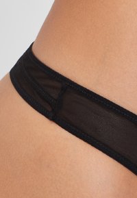 Musta läpinäkyvä verkkopanta, jossa on leveä elastinen vyötärönauha. Yksinkertainen muotoilu ja sileä rakenne korostavat tarkkaa ompelua ja saumoja.