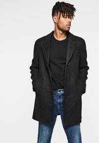 Abrigo de mezcla de lana negro con solapas en pico, dos botones y dos bolsillos frontales, usado sobre un suéter negro de cuello redondo y jeans de mezclilla.