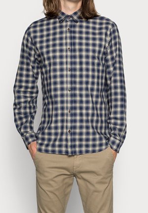 Homme portant une chemise à carreaux bleu marine et beige boutonnée avec un pantalon kaki, debout les mains dans les poches devant un fond uni.