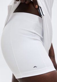 Zbliżenie na osobę noszącą obcisłe białe sportowe spodenki z małym czarnym logo blisko brzegu, podnoszącą białą koszulkę, aby odsłonić pas biodrowy.