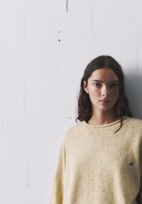 Pulover tricotat de culoare galben deschis, cu un finisaj texturat, decolteu larg și logo brodat pe partea stângă. Modelul stă în fața unei pereți albe.