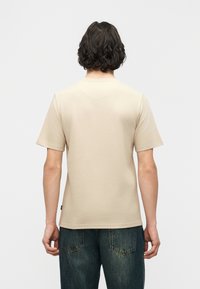 Άτομο με κοντά σγουρά μαλλιά φοράει απλό μπεζ κοντομάνικο πουκάμισο και σκούρο μπλε τζιν, στέκεται με την πλάτη σε απλό φόντο.