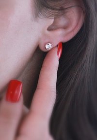 Orecchino a stella in argento con una pietra preziosa trasparente, vicino all'orecchio, mano con smalto rosso che tocca l'orecchino, capelli morbidi visibili.