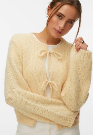 Femme portant un cardigan en tricot doux de couleur beige clair avec deux nœuds noués à l'avant, associé à un pantalon blanc et un haut blanc en dessous.