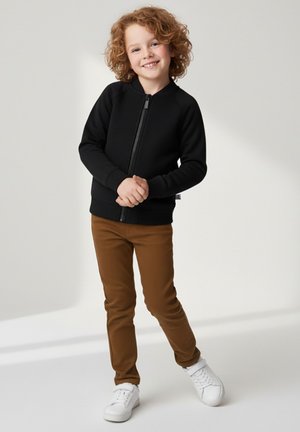 Czarna bluza na zamek z gładką fakturą, zestawiona z dopasowanymi brązowymi spodniami i białymi tenisówkami, prezentująca funkcjonalny i casualowy design.