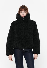 Zwarte, fluffy jas met een hoge kraag en ritssluiting; brede mouwen en textiel met een structuur. Gepaard met donkere jeans.