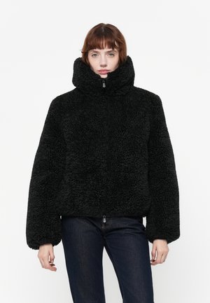 Zwarte, fluffy jas met een hoge kraag en ritssluiting; brede mouwen en textiel met een structuur. Gepaard met donkere jeans.