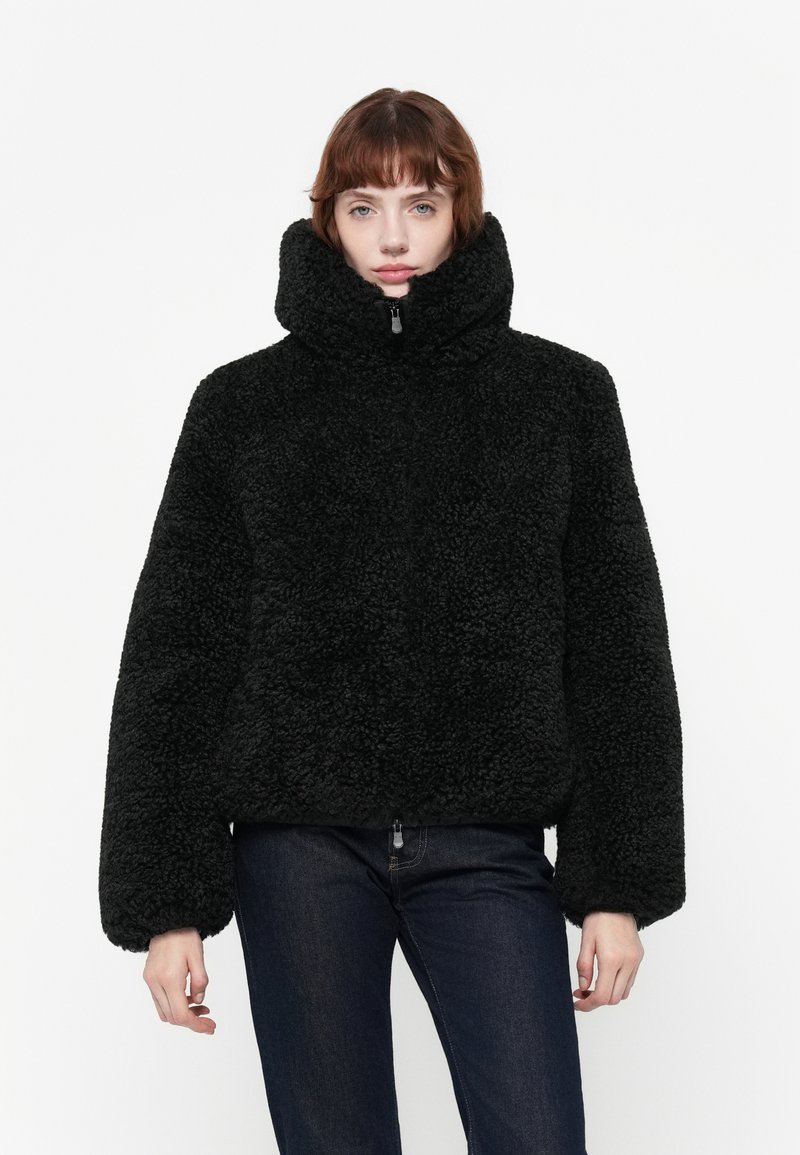Zwarte, fluffy jas met een hoge kraag en ritssluiting; brede mouwen en textiel met een structuur. Gepaard met donkere jeans.