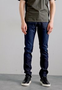 Tamsiai mėlynos džinsinės kelnės slim fit stiliaus, pagamintos iš džinsinio audinio. Derinamos su juodais sportbačiais ir žaliu marškinėliu, stovintis prieš šviesų foną.