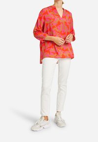Langsleeve, roze blouse met abstracte patronen in oranje en fuchsia; v-hals; losse pasvorm; gecombineerd met witte broek en lichte sneakers.
