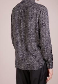 Camicia nera a maniche lunghe con un motivo geometrico bianco che presenta motivi di teschi. Il tessuto ha una texture liscia e un design classico del colletto.