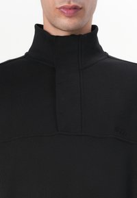 Maglione nero con colletto alto, dotato di chiusura con zip e dettagli cuciti. Il materiale appare liscio con vestibilità slim e logo del brand.