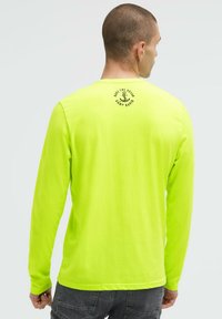 Langärmeliges, leuchtend neon-gelbes Shirt aus weichem Stoff, mit einem kreisförmigen schwarzen Grafikmotiv und Text auf der Rückseite sowie einem Rundhalsausschnitt.