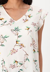 Nainen, jolla on yllään hihaton valkoinen paita, jossa on värikäs kukkakuosi ja V-pääntie, joka paljastaa ylävartalon ja hartiat.