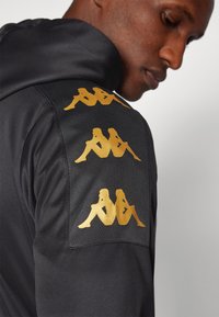 Black hooded jacket featuring three gold logos on the shoulder, made from smooth fabric with a textured panel on top.