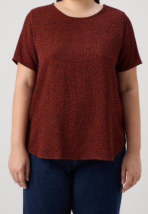 Person trägt ein kurzärmeliges, rostfarbenes Hemd mit kleinem schwarzem Leopardenmuster und dunkelblaue Jeans.