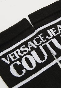 Črne pletene nogavice z belim debelim besedilom "VERSACE JEANS COUTURE" in belimi vodoravnimi črtami blizu zgornjih robov.