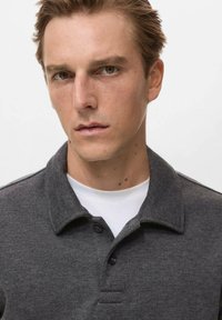 Dunkelgraues Poloshirt aus strukturiertem Material, mit Kragen und drei Knöpfen, über einem weißen Unterhemd getragen.