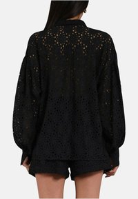 Camicia di pizzo nera con maniche lunghe e collo. Dettagli floreali intricati con ritagli. Abbinata a pantaloni corti in pizzo coordinati.