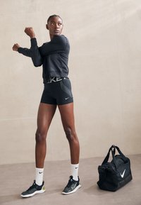 Fekete Nike pulóver és rövidnadrág logóval, texturált zoknik és fekete sportcipők. Fekete tornazsák a padlón, fehér logóval. Simább háttér.