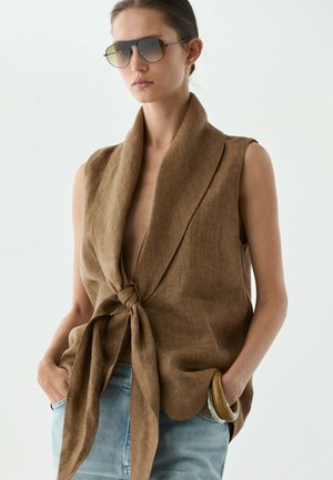 Kvinde iført brun ærmeløs top med bindebånd foran, lyseblå jeans, stor armring på håndleddet og mørke solbriller, der står mod en ensfarvet baggrund.