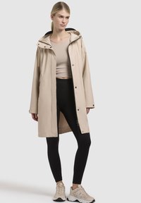 Beige waterdichte jas met een capuchon, drukknopen aan de voorkant en zijzakken, gecombineerd met zwarte leggings en beige sportschoenen.